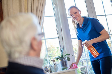 This is as photographic image of a young female with a blond ponytail and wearing a blue polo shirt that is spray cleaning a tasble while tallking to an elderly gentleman with white hair, light blue shirt, and dark blue sweater in the foregroung.