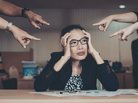 Woman sitting at a desk looking stressed while fingers point at her from different directions.