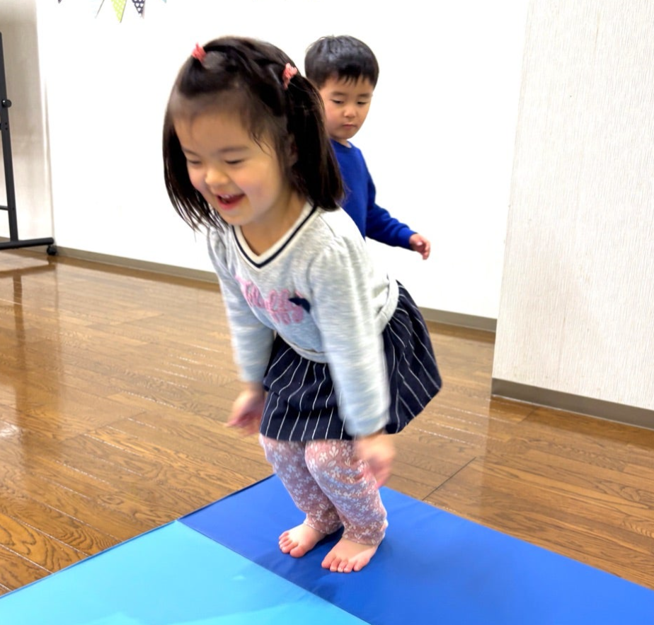 室内で女の子が青いマットの上でジャンプし、後ろに男の子が見守る。女の子は笑顔で、背景は白い壁で明るい雰囲気。