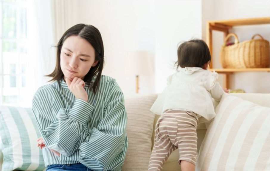 ストライプのシャツを着た女性が、ソファで遊ぶ子供を見つめて考えている。背景にバスケットが置かれた棚が見える。