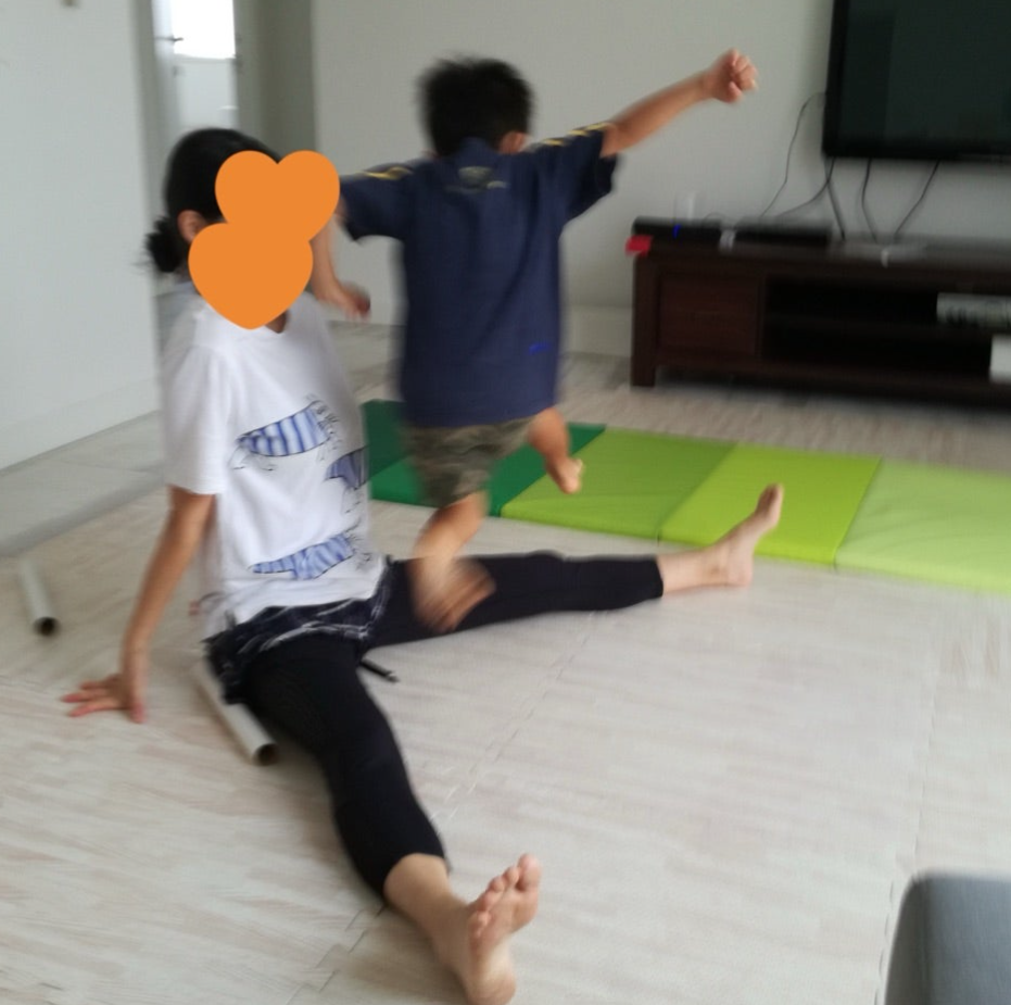 白いTシャツの大人が床に座り、子供が飛び跳ねる様子。背景にテレビとカラフルなマット。明るい室内で楽しそうな雰囲気。