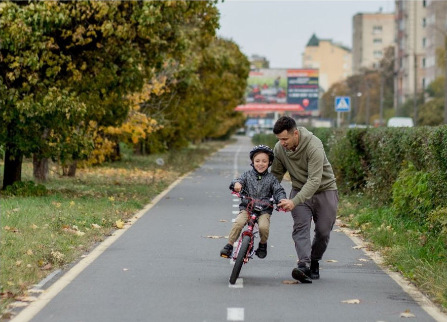 父親が公園の道で息子に自転車の乗り方を教えている。息子はヘルメットをかぶり笑顔。秋の葉が彩る背景。楽しげな雰囲気。