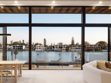 Modern living room with wooden dining table and white sofa facing large windows overlooking a serene waterfront and cityscape at sunset.