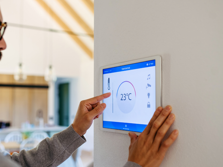 A person adjusts a wall-mounted digital thermostat showing 23°C. The background features a modern interior with warm lighting.