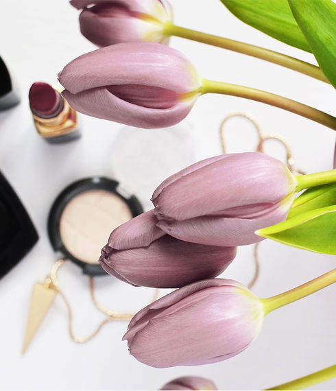 Lavender Tulips with green folage in the foreground. Out of focus, deep red lipstick, a pink eyeshadow, and gold necklace with triagular pendent in the background.