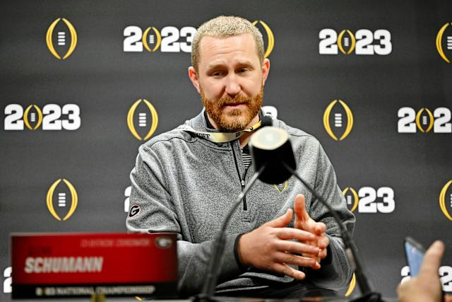 Man in a gray sweater speaking at a press conference, microphones visible. Background has "2023" and a football emblem. Mood is attentive.