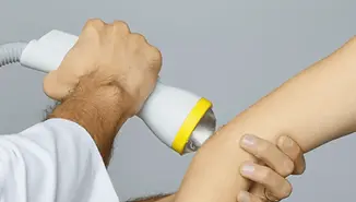 a softwave machine being held by a white lab coat arm and being applied to a persons elbow with ultrasound gel on the elbow.