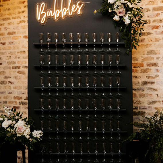 Large vertical champagne glasses on black background with the words Bubbles in cursive lit up in orange and flowers on the top right