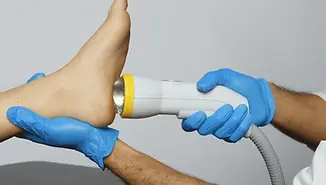 a softwave machine being held by a white lab coat arm and being applied to a persons foot with ultrasound gel on the foot.