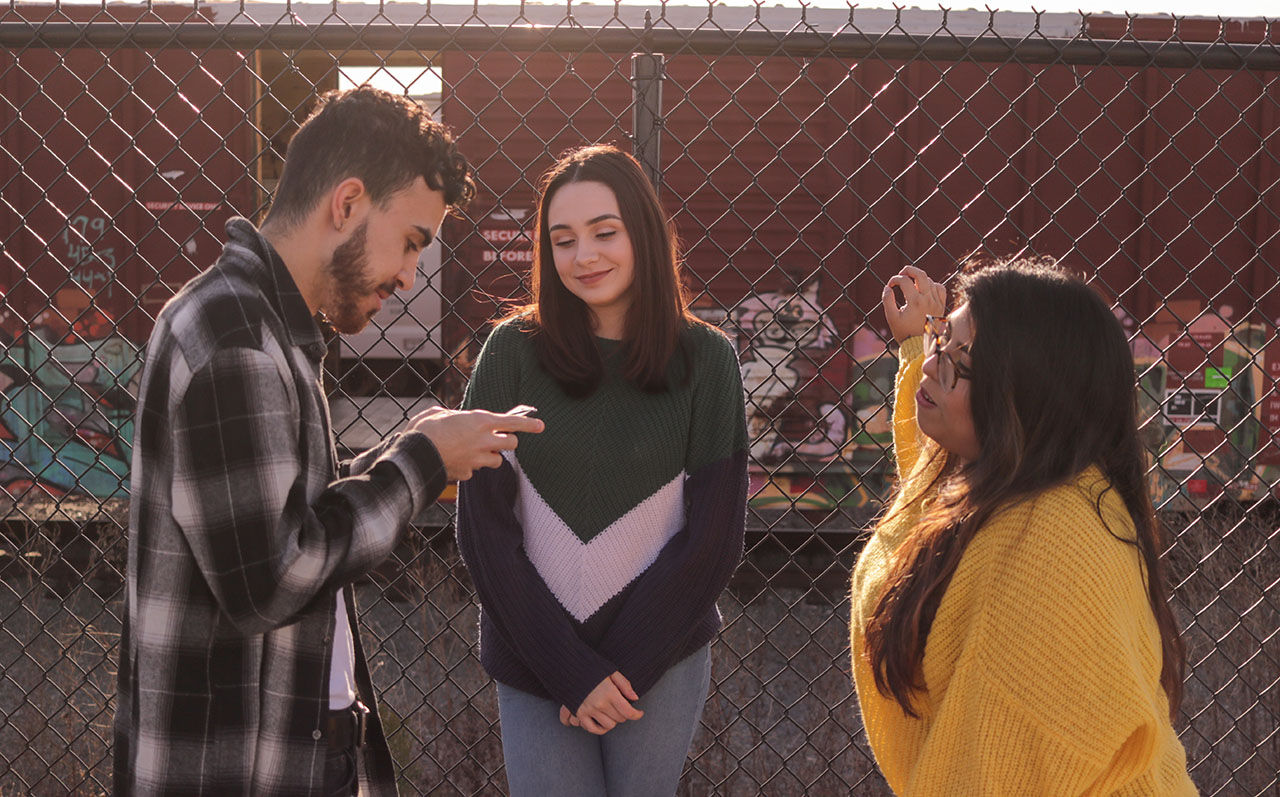 Two women and a man stand in front of a fence and talk.