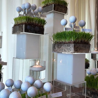 Golf-inspired escort table design for a mitzvah celebration featuring seating cards displayed on golf balls, modern acrylic risers, and lush grass accents.