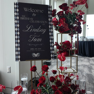 Black and white diamond-pattern welcome sign for Lindsay and Tom’s surprise birthday celebration, surrounded by lush red roses, anthuriums, and floating candles at Vue on 50, designed by Philadelphia Event Decor.
