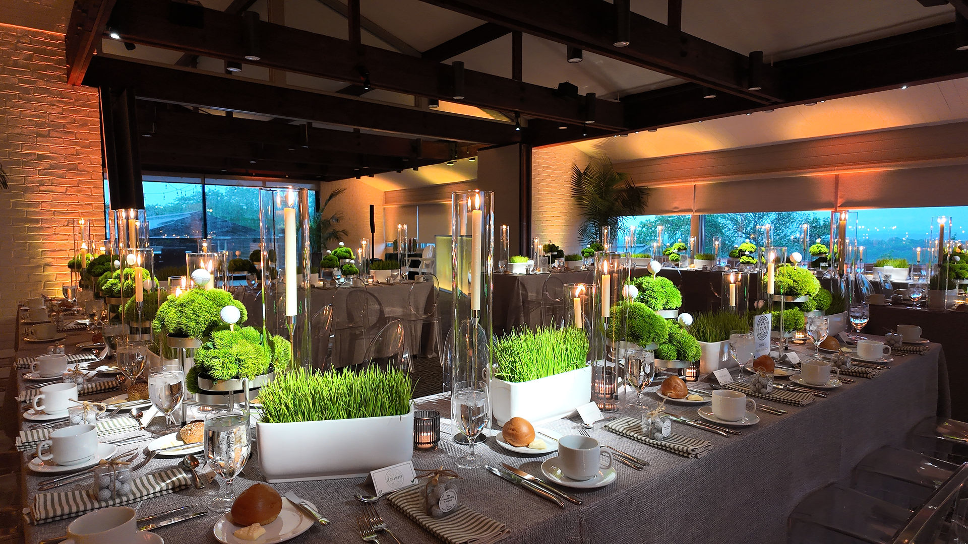 Modern bar mitzvah reception tablescape featuring minimalist greenery centerpieces, tall glass candle holders, and neutral place settings in a contemporary indoor venue.