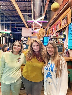 Three people stand closely together posing for a photo inside a warmly lit bar or brewery. The person in the center wears a yellow sweater, while the two people beside them wear light-colored sweatshirts—one with a snowman graphic and the other with a Detroit Lions logo. Behind them are bar shelves, taps, colorful neon lights, and festive hanging ornaments.