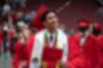 A young man dressed in a cap and gown with an 'AVID' stole smiles and waves while displaying diploma
