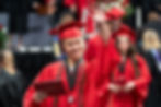 A young man dressed in a cap and gown waving and smiling while holding diploma