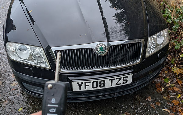 A hand holding a Skoda car key fob in front of a parked black Skoda.