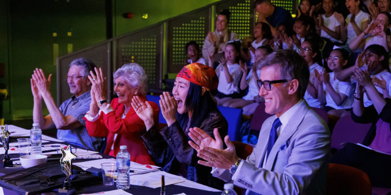 Judges and audience clap and cheer in a dim auditorium, smiling under purple-green lights at a live event.