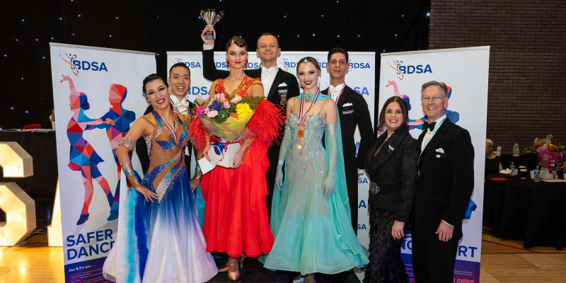 Award-winning ballroom dancers pose onstage with a trophy and bouquet at a BDSA event, flanked by judges and banners.