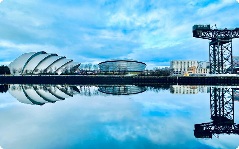Glasgow waterfront skyline with a crane, domed buildings, and mirror-like reflections under a blue cloudy sky.