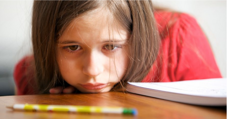 Niña desanimada apoyando su mentón en un escritorio, con su cuaderno a un costado y mirando fijamente y con mirada triste un lápiz que tiene enfrente apoyado en el escritorio. No se le ven los brazos lo que refuerza la sensación de desaliento.