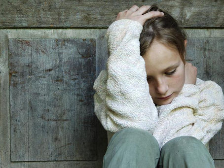 Niña sentada con su cabeza entre una mano y la otra mano sobre el cuello, contra una pared en posición de tristeza y abatimiento.