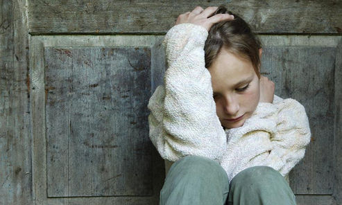 Niña sentada con su cabeza entre una mano y la otra mano sobre el cuello, contra una pared en posición de tristeza y abatimiento.