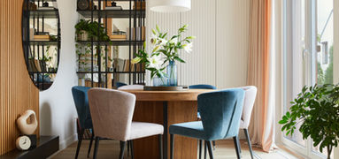 Bright dining area with a round wooden table, upholstered chairs in soft neutral and blue tones, open shelving with plants, and natural light from a large window.