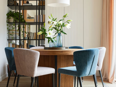 Bright dining area with a round wooden table, upholstered chairs in soft neutral and blue tones, open shelving with plants, and natural light from a large window.