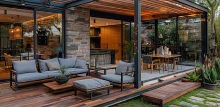 Modern open-air patio with gray sofas, wood deck, glass walls, warm lights, and a cozy dining area beside lush plants.