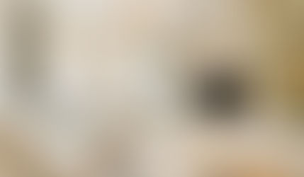 Transitional living room with beige sofa, chair, TV on white shelves, and a marble table on floral rug. Large windows and chandelier overhead.