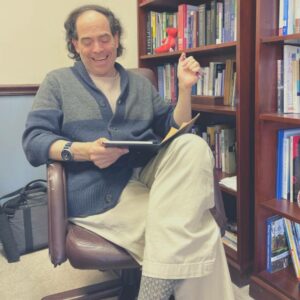 man in chair reading a book in front of bookshelf