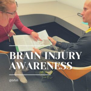 2 people sit across from each other, one points to a page in an open book