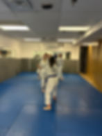 Martial arts class in session. Five people in white uniforms practice on blue mats. Mirrors reflect the scene. Posters on walls enhance focus.