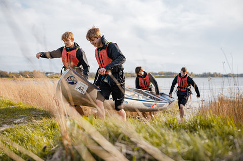 Gruppe af unge personer iført redningsveste bærer en kano ved en sø i naturskønt landskab. Perfekt til outdoor- og eventfotografi, der fremhæver teamwork og adventure-aktiviteter.