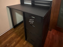 Black desk with three drawers and shelves, placed in a room with wooden flooring. Branding text "my-desk.co" visible on the drawers.