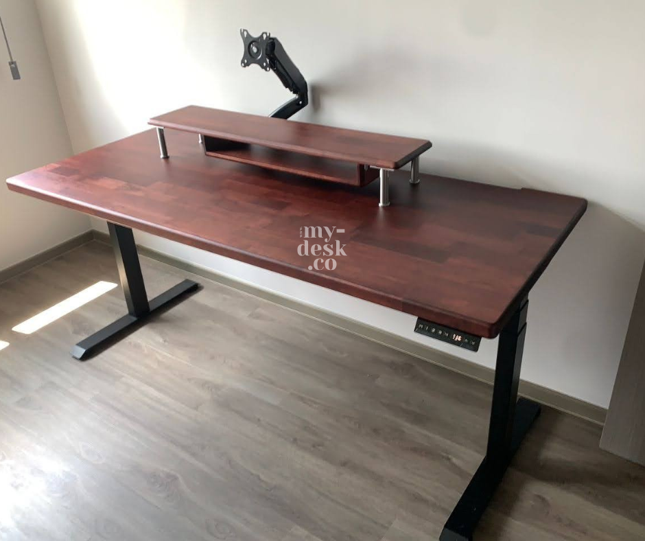 Wooden desk with adjustable legs and a monitor stand against a light wall. Floor is wooden. Sunlight casts shadows. "my-desk.co" text visible.