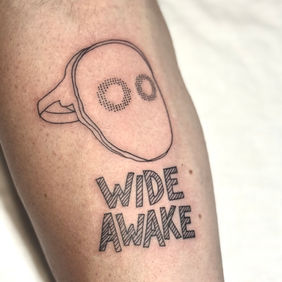 Tatouage fineline masque minimaliste et lettrage sur avant-bras au salon La Fratrie à Nice.