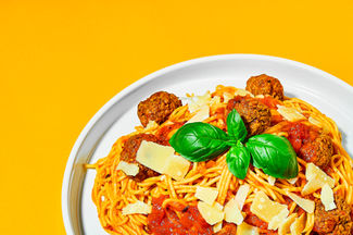 food photography of spaghetti meat balls plated on a white plate garnished with basil and parmesan shavings set on a bold orange backdrop with a tight crop, Yorkshire 