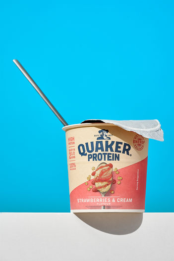 commercial food phot of quakers protein, set on a white foreground with a light blue background, quakers lid is hard open and a spoon showing out of the oats pot 