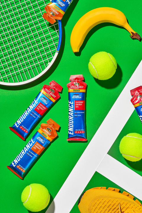 Applied Nutrition Endurance energy gels with tennis racket, tennis balls, banana and trainer on a vivid green court background — sports nutrition product photography UK