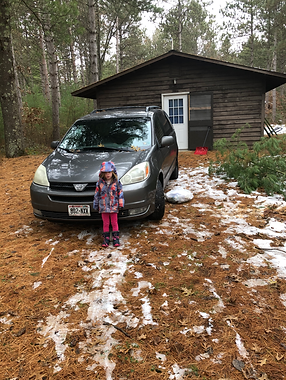 A little girl stands in front of a minivan in front of a cabin and Camp Helen Brachman. Small piles of snow are seen on top of the dirt road