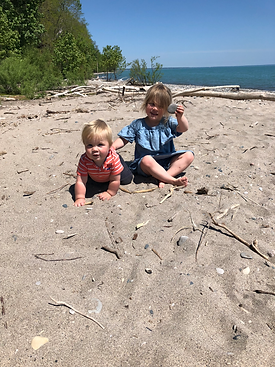 a sister and her baby brother collect rocks at the beach. the girl is sitting in the sand holding up her rock and the baby is crawling on the sand. it is a sunny day with clear skies