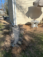 A completed water feature at the end of a white downspout of an old farmhouse. The water feature is lined with big rocks and filled with river pebbles and river rocks