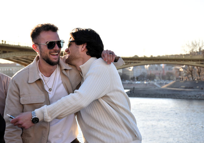 Two laughing men embracing by a river and bridge.