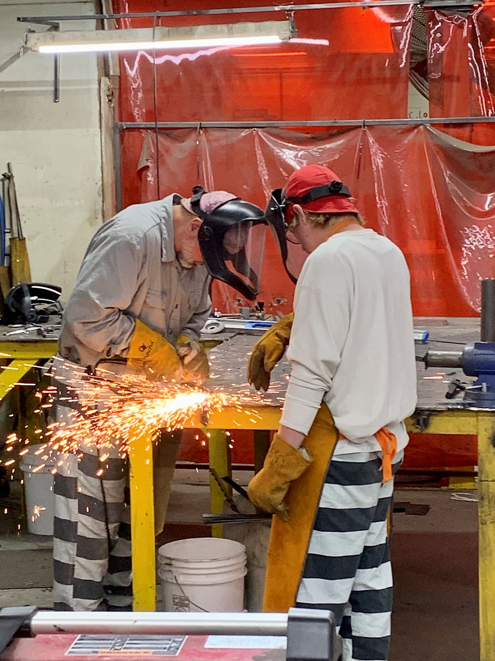 An older gentleman in black an white stripes wearing safety gear is welding with a younger guy in black and white stripes and safety gear watching him. This is mentoring in action.