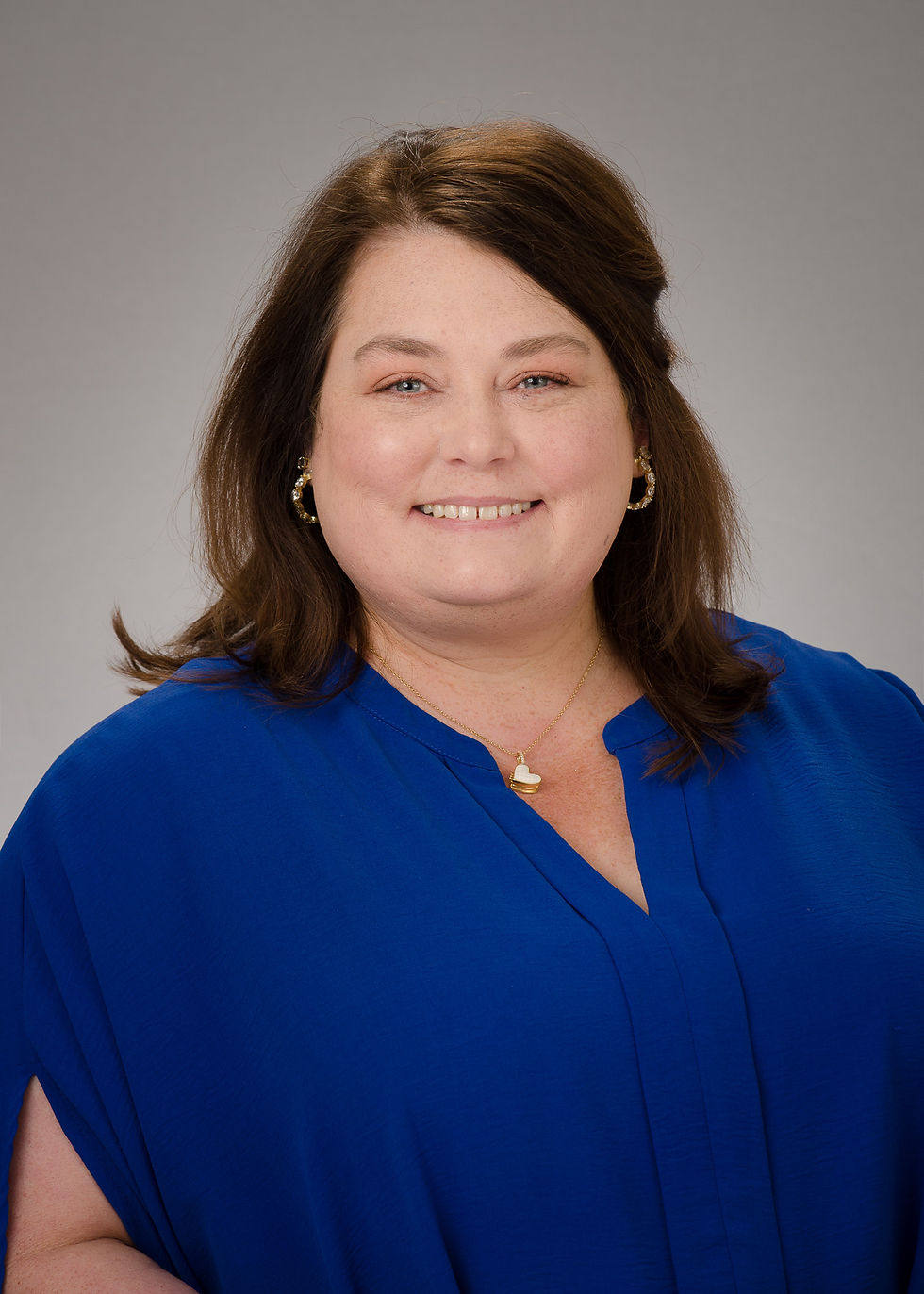 A woman with brown shoulder length hair with one side pulled back is smiling. She wears a royal blue shirt with a gold chain necklace and gold earrings.