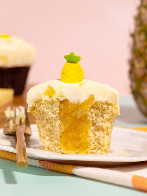Cupcake with pineapple filling and frosting, topped with a pineapple decoration. Set on a plate with a fork, against a pastel backdrop.