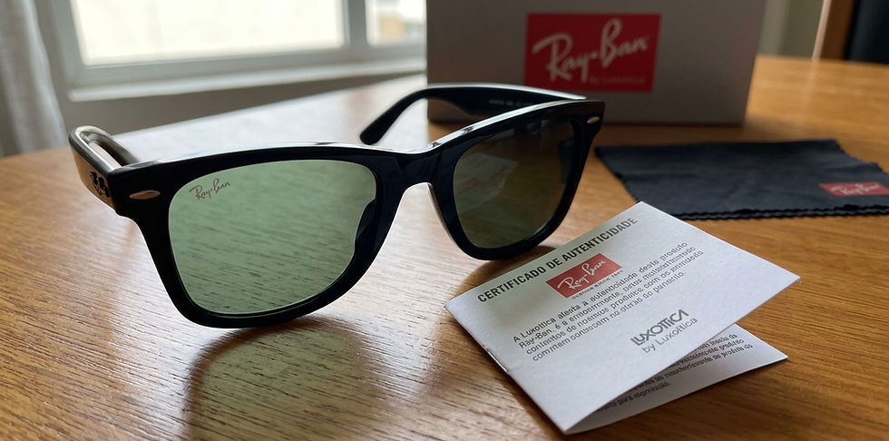 Óculos de sol Ray-Ban preto sobre mesa de madeira. Certificado de autenticidade e estojo no fundo. Luz suave da janela. Emblema visível.