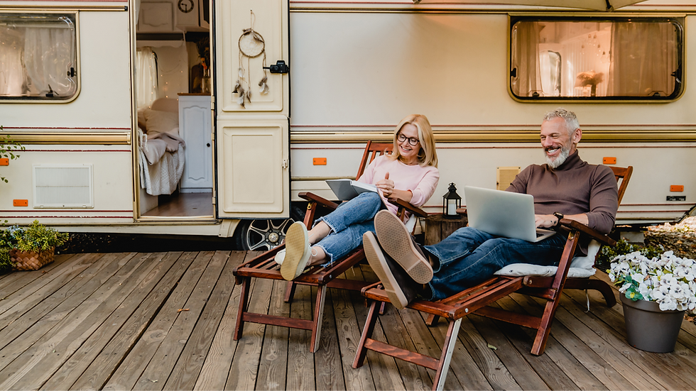 Couple souriant devant une caravane, assis sur des chaises longues avec des ordinateurs portables, sur une terrasse en bois.
